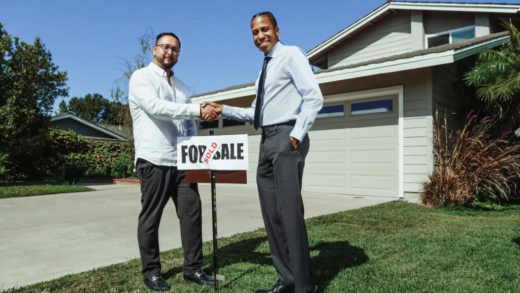 Two men shake hands in front of a new home with a sold sign, symbolizing a successful real estate de